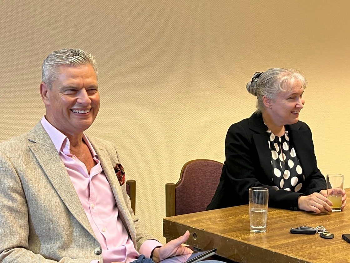 heller Tapetenhintergrund, links vom Tischausschnitt sitzend Ingo Hoppe, rechts davon Pamela Pabst, beide in unterschiedliche Richtungen lächelnd, auf dem Tisch Getränke, Kfz-Schlüssel